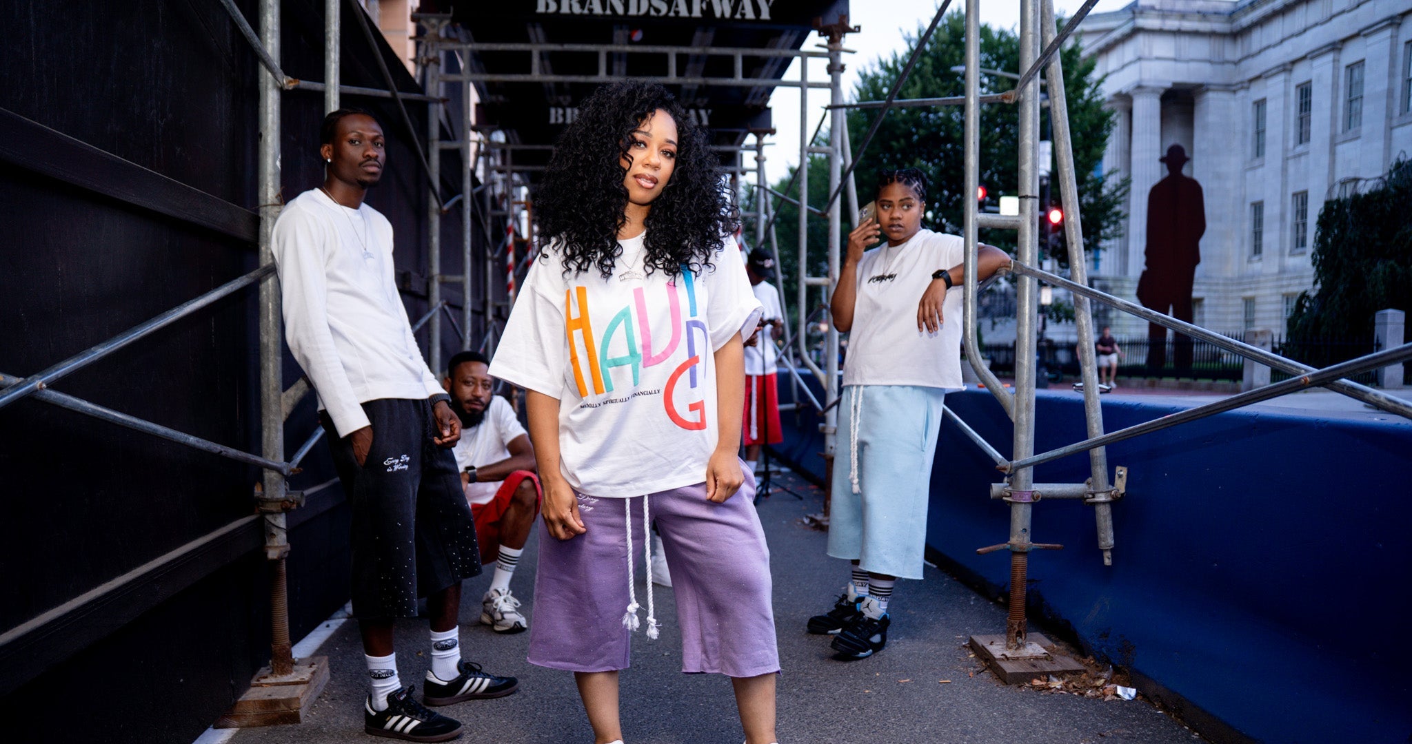 A group of models posing on a city street under scaffolding, featuring a central model wearing a white graphic t-shirt with colorful 'HAVING' lettering and lavender long shorts.